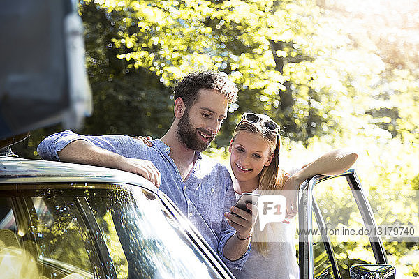 Smiling young couple with cell phone at car in forest