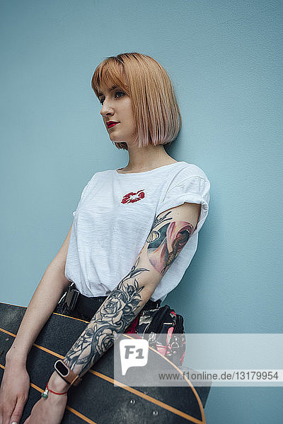 Coole junge Frau hält Carver-Skateboard in der Hand und steht an türkisfarbener Wand