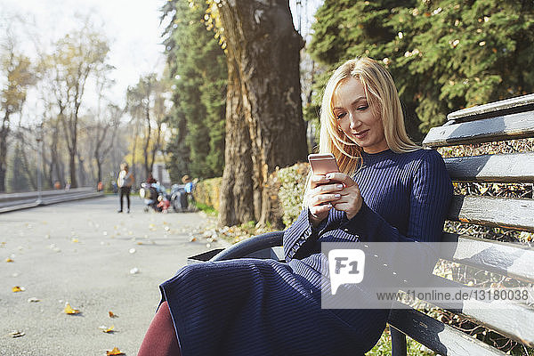 Porträt einer blonden Frau  die auf einer Bank im herbstlichen Stadtpark sitzt und ein Smartphone benutzt