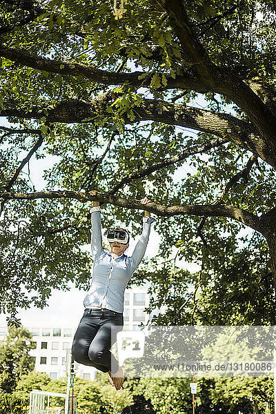 Frau im Stadtpark  die am Ast eines Baumes hängt und eine VR-Brille trägt