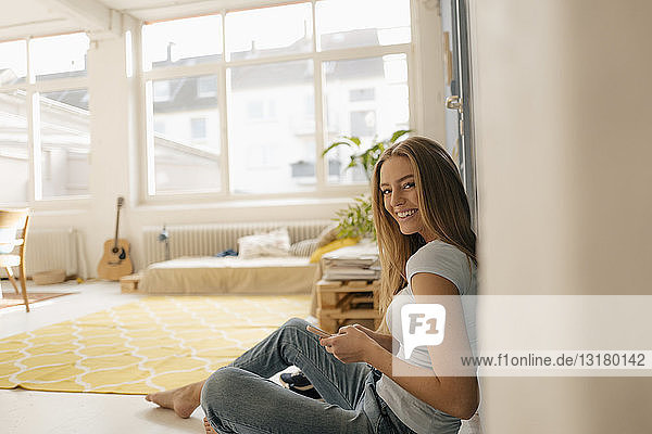 Porträt einer lächelnden jungen Frau  die in ihrem Loft auf dem Boden sitzt und ein Smartphone benutzt