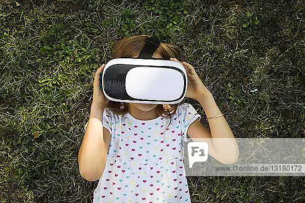 Kleines Mädchen mit Virtual-Reality-Brille auf der Wiese im Garten liegend