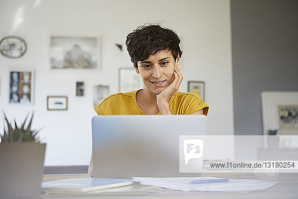 Porträt einer lächelnden Frau,  die zu Hause am Tisch sitzt und einen Laptop benutzt