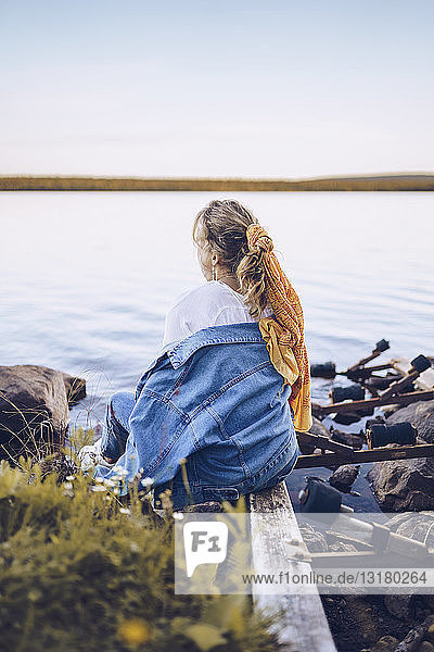 Schweden  Lappland  Rückenansicht einer jungen Frau  die am Wasser sitzt und in die Ferne schaut