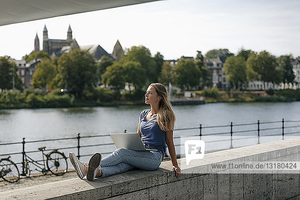 Netherlands  Maastricht  young woman sitting on a wall at the riverside with laptop