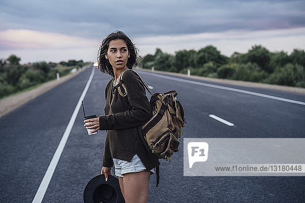Portrait of young hitchhiking woman with backpack and beverage standing on lane