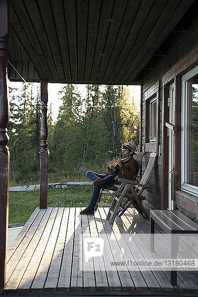 Junger Mann sitzt auf der Veranda eines Holzhauses und spielt Ukulele