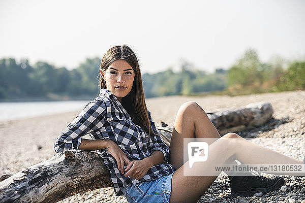 Junge Frau sitzt am Kieselsteinufer am Baumstamm
