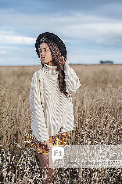 Porträt einer jungen Frau mit Hut und übergroßem Rollkragenpullover im Maisfeld stehend