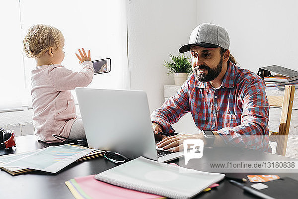 Vater arbeitet von zu Hause aus  benutzt einen Laptop  während seine Tochter auf dem Schreibtisch sitzt und spielt