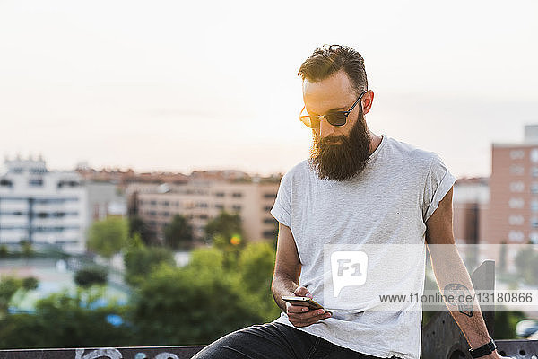 Cooler bärtiger junger Mann mit Sonnenbrille beim Handy-Check