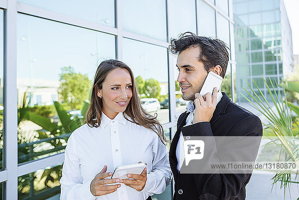 Smiling businesswoman and businessman with tablet and cell phone outside office building