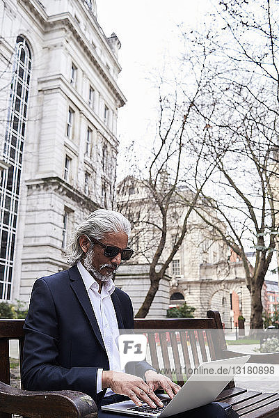 UK  London  senior businessman sitting on bench outdoors working on laptop