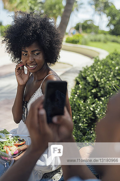 USA  Florida  Miami Beach  junger Mann beim Fotografieren einer Freundin  die in einem Park einen Salat isst