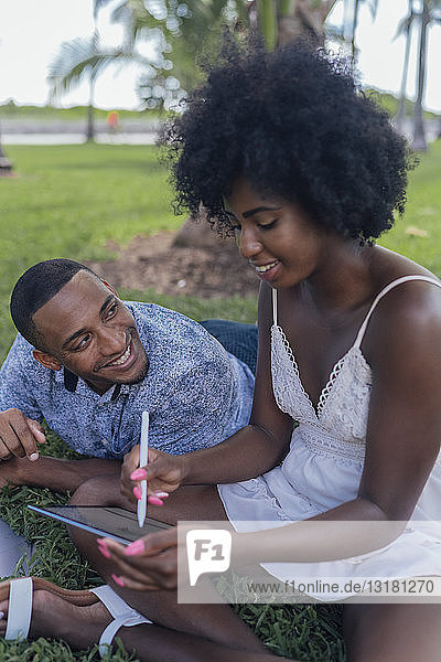 USA  Florida  Miami Beach  lächelnder junger Mann  der seine Freundin mit einer Tablette auf einem Rasen in einem Park ansieht