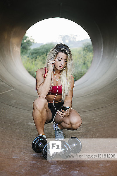 Athletische Frau  die mit Hanteln und Kopfhörern in einer Röhre hockt und auf ihr Handy schaut
