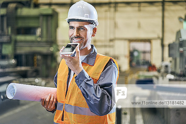 Smiling man holding blueprints and using cell phone in factory