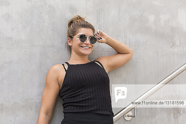 Porträt einer lächelnden jungen Frau mit Sonnenbrille  die an einer Betonwand lehnt