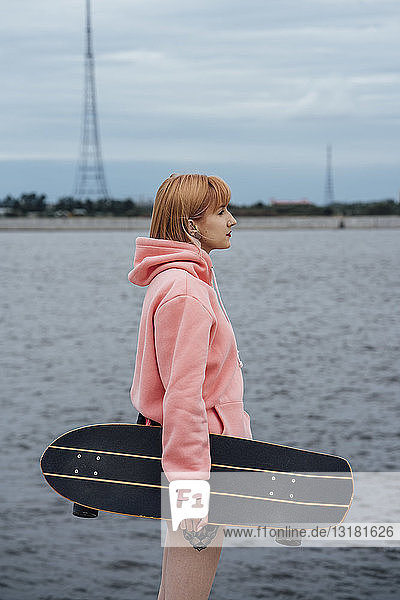 Junge Frau hält Carver Skateboard in der Hand und steht am Flussufer