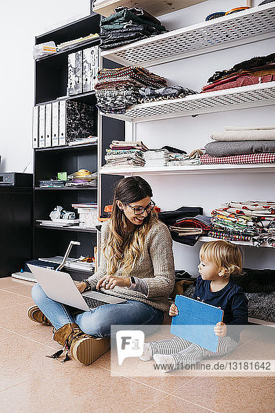 Mutter und kleine Tochter sitzen zu Hause mit Laptop und Tablett auf dem Boden
