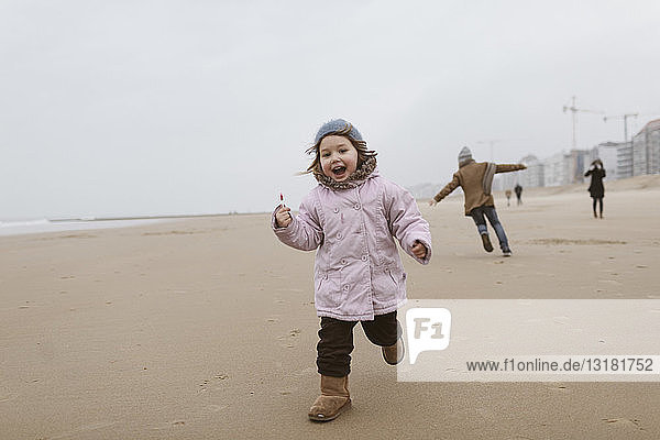 Happy little girl with lolly running on the beach in winter while her brother playing in the background