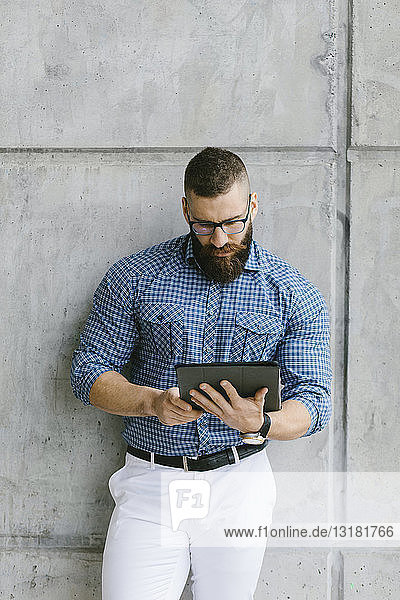 Bärtiger Hipster-Geschäftsmann mit Brille und kariertem Hemd mit Tablette an die Wand gelehnt