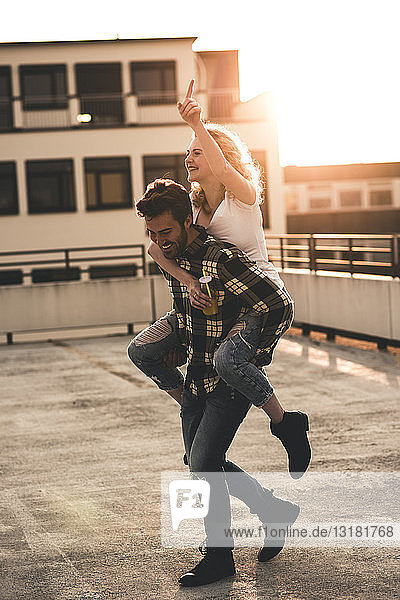 Glücklicher junger Mann  der seine Freundin bei Sonnenuntergang huckepack auf der Dachterrasse mitnimmt