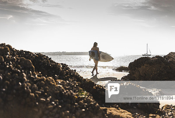 Frankreich,  Bretagne,  junge Frau trägt Surfbrett an einem felsigen Strand am Meer