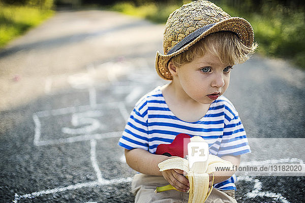 Porträt eines Kleinkindes mit Banane  das auf der Straße sitzt und etwas beobachtet