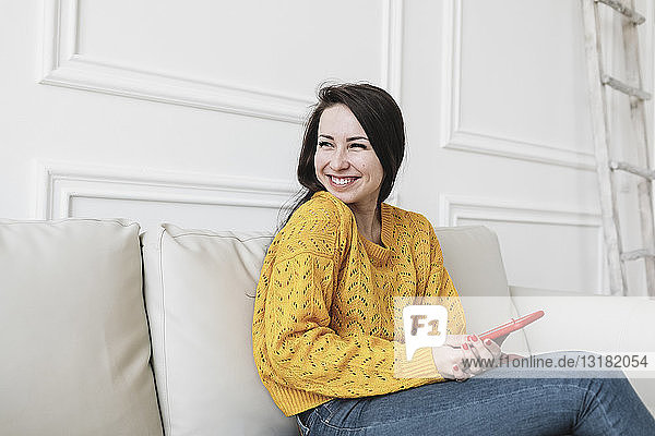 Porträt einer genossenen jungen Frau mit Smartphone  die in ihrem neuen Zuhause auf der Couch sitzt und in die Ferne schaut
