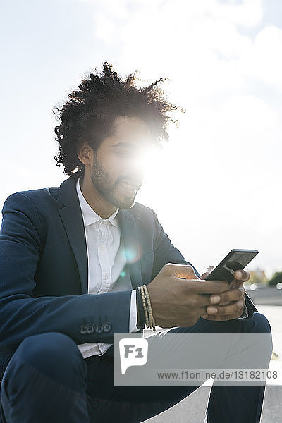 Junger Geschäftsmann sitzt im Freien bei Sonnenschein und benutzt sein Handy