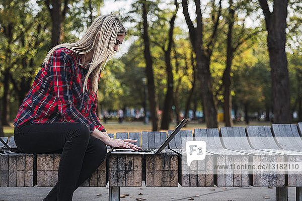 Junge Frau sitzt auf Bank im Freien und arbeitet am Laptop