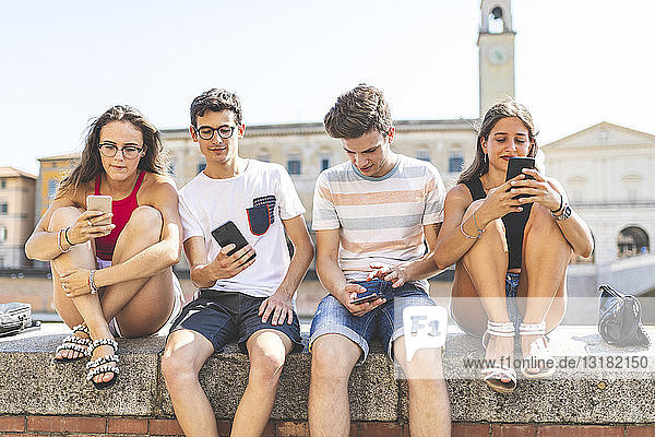 Italien  Pisa  Gruppe von vier Freunden  die gemeinsam mit Mobiltelefonen an einer Wand sitzen