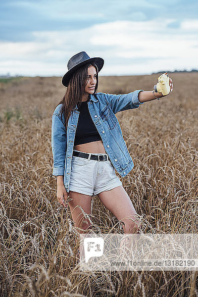 Junge Frau mit Hut und Jeansjacke  die mit Kamera in einem Maisfeld Selbstporträt macht