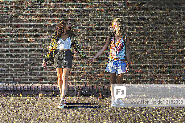 Zwei junge Frauen halten sich vor einer Ziegelmauer an den Händen