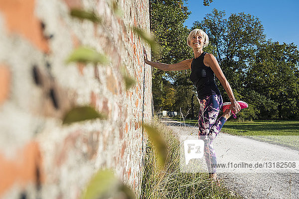 Lächelnde sportliche ältere Frau  die sich an einer Ziegelmauer ausdehnt