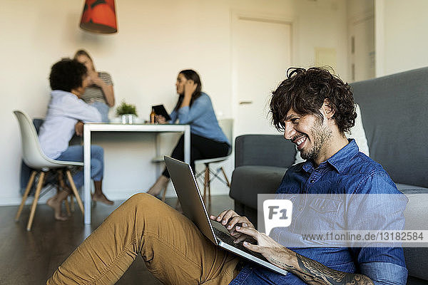 Lächelnder Mann sitzt auf dem Boden und benutzt einen Laptop mit Freunden im Hintergrund