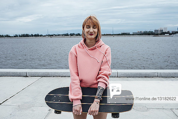 Lächelnde junge Frau hält Schnitzerin Skateboard stehend am Flussufer