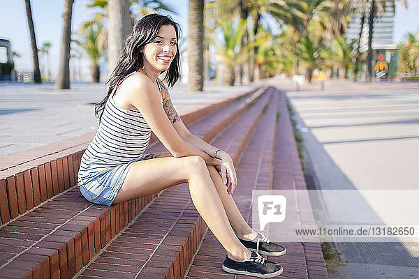 Spanien  Barcelona  Porträt einer lächelnden jungen Frau mit Nasenpiercing und Tätowierungen  die im Sonnenlicht auf Stufen sitzt
