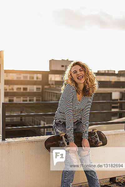 Portrait of cheeky young woman with skateboard on roof terrace at sunset