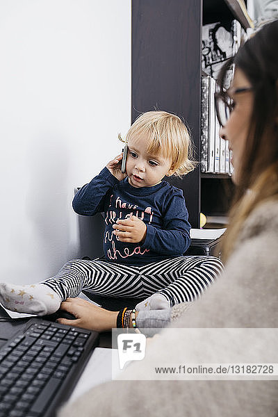 Mother working at home with little daughter sitting on desk holding cell phone