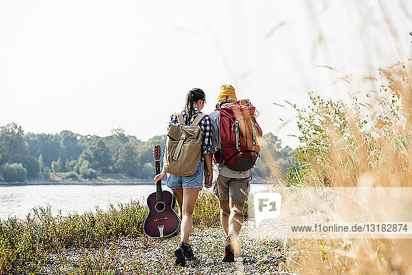 Rückansicht eines jungen Paares mit Rucksäcken und Gitarre beim Spaziergang am Flussufer