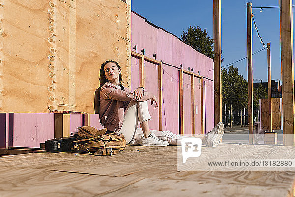 Entspannte junge Frau sitzt auf Podest neben Gitarrenkoffer und Tasche