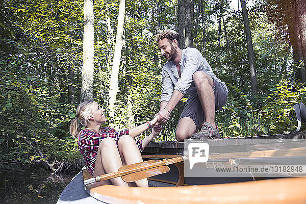 Lächelnder junger Mann hilft seiner Freundin in einem Waldbach beim Aussteigen aus dem Kanu
