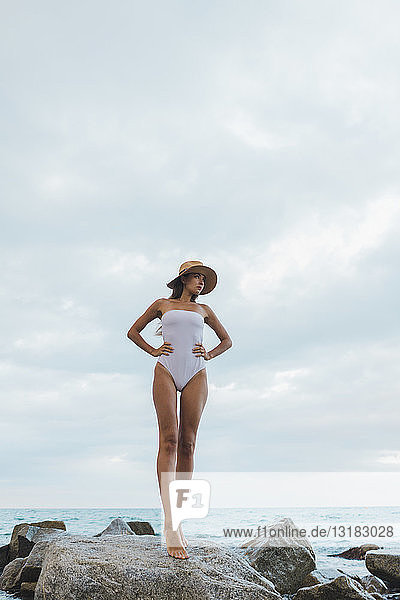 Beautiful young woman wearing swimsuit and hat standing on rock in the sea