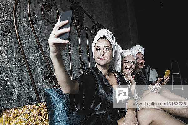 Three women with towels around her heads on bed taking a selfie
