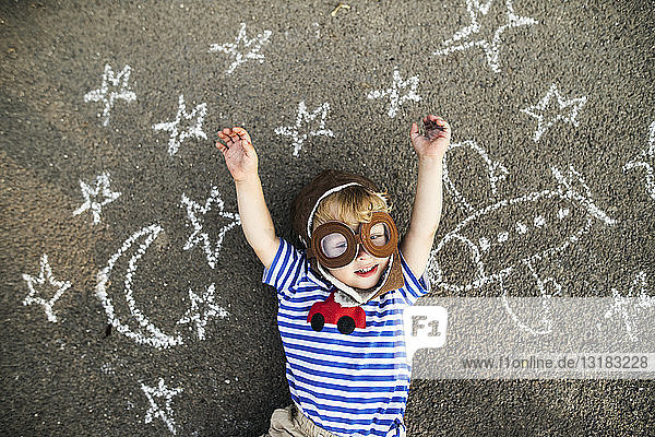 Porträt eines lächelnden Kleinkindes mit Pilotenhut und Schutzbrille auf Asphalt liegend  bemalt mit Flugzeug  Mond und Sternen