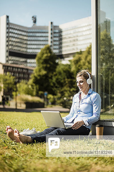 Lächelnde Frau mit Kopfhörern sitzt mit Laptop auf städtischer Wiese
