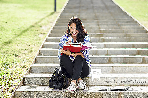 Lächelnder Student sitzt auf einer Treppe im Freien und macht Notizen in einem Notizbuch