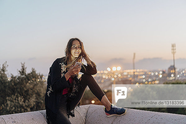 Spanien  Barcelona  Montjuic  lächelnde junge Frau  die in der Abenddämmerung mit ihrem Handy an einer Wand sitzt